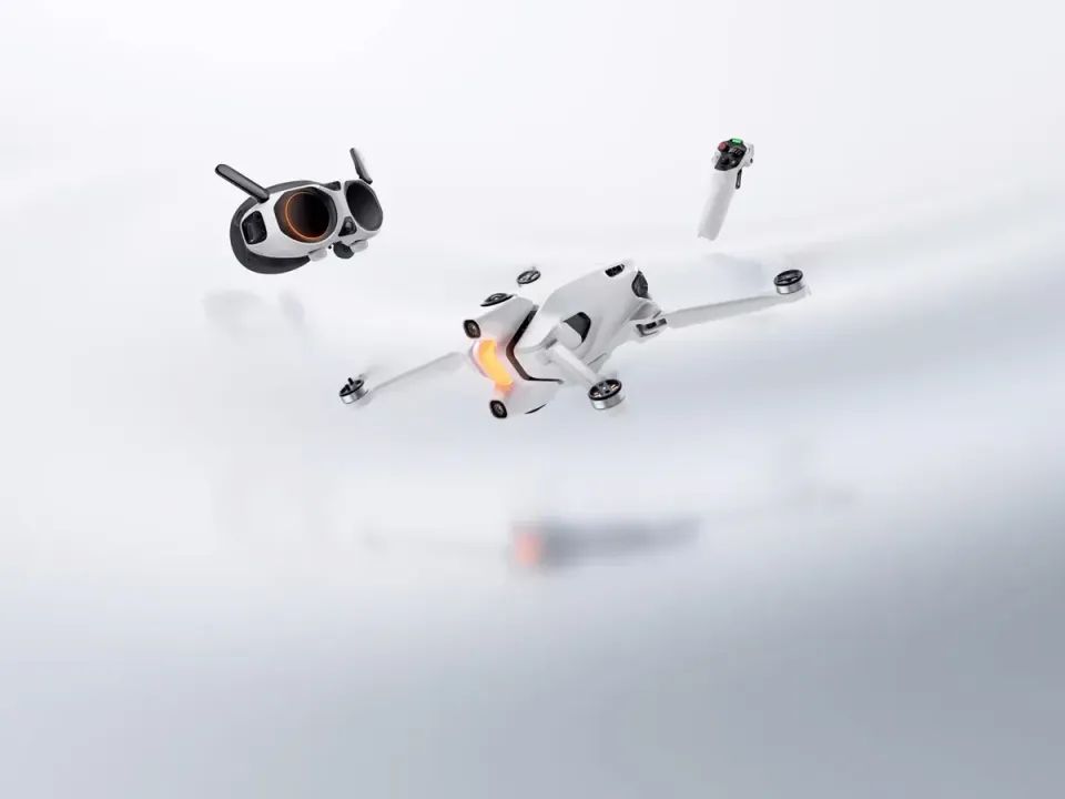 White Antigravity A1 quadcopter in flight beside its white goggles and a single-hand grip controller against a pale grey studio background