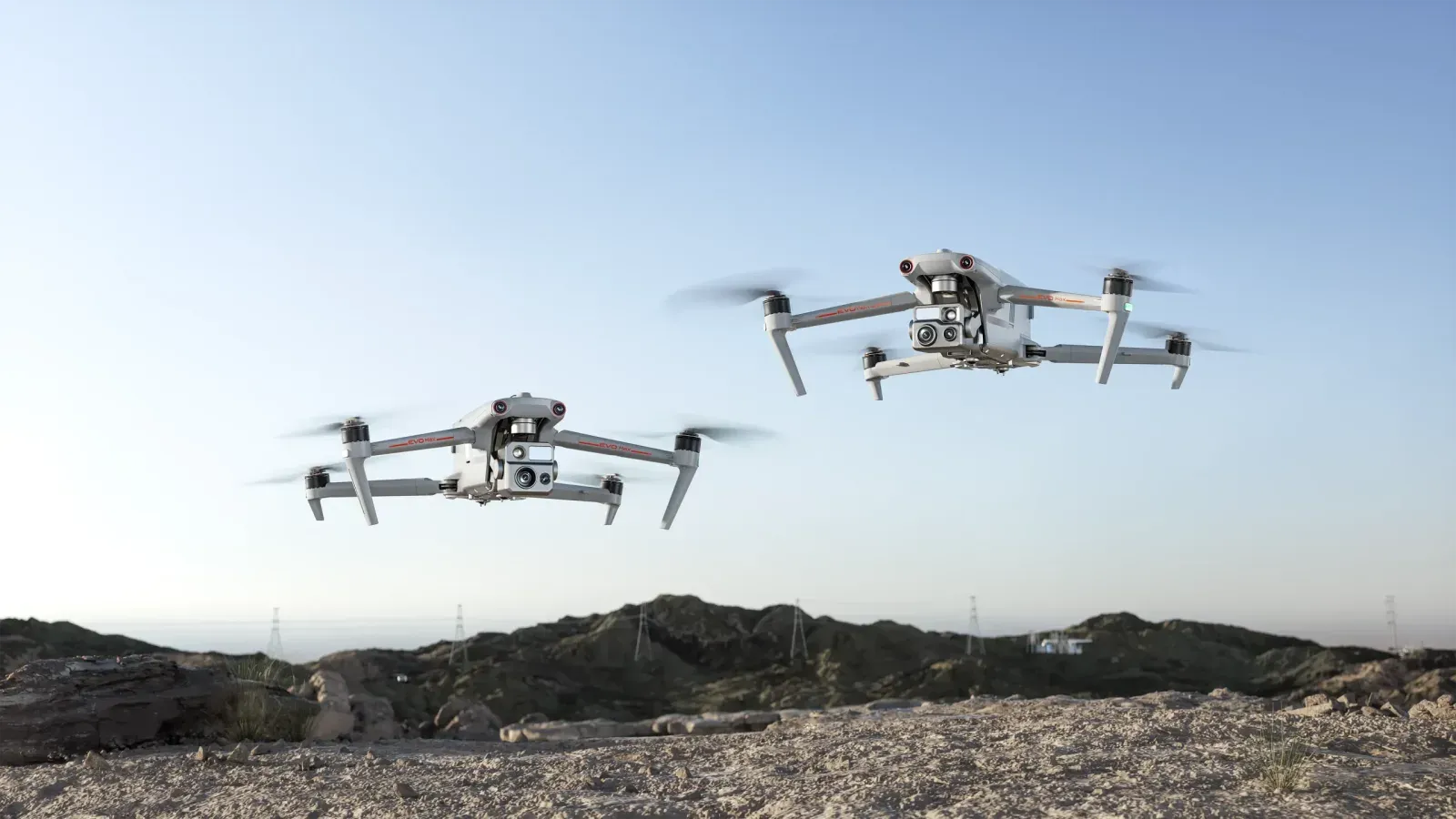 Two grey Autel EVO Max quadcopters with orange trim hovering over a rocky ridge with power pylons behind