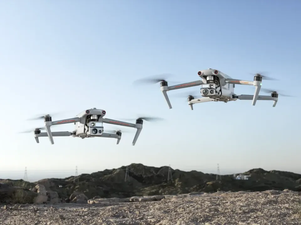Two grey Autel EVO Max quadcopters with orange trim hovering over a rocky ridge with power pylons behind