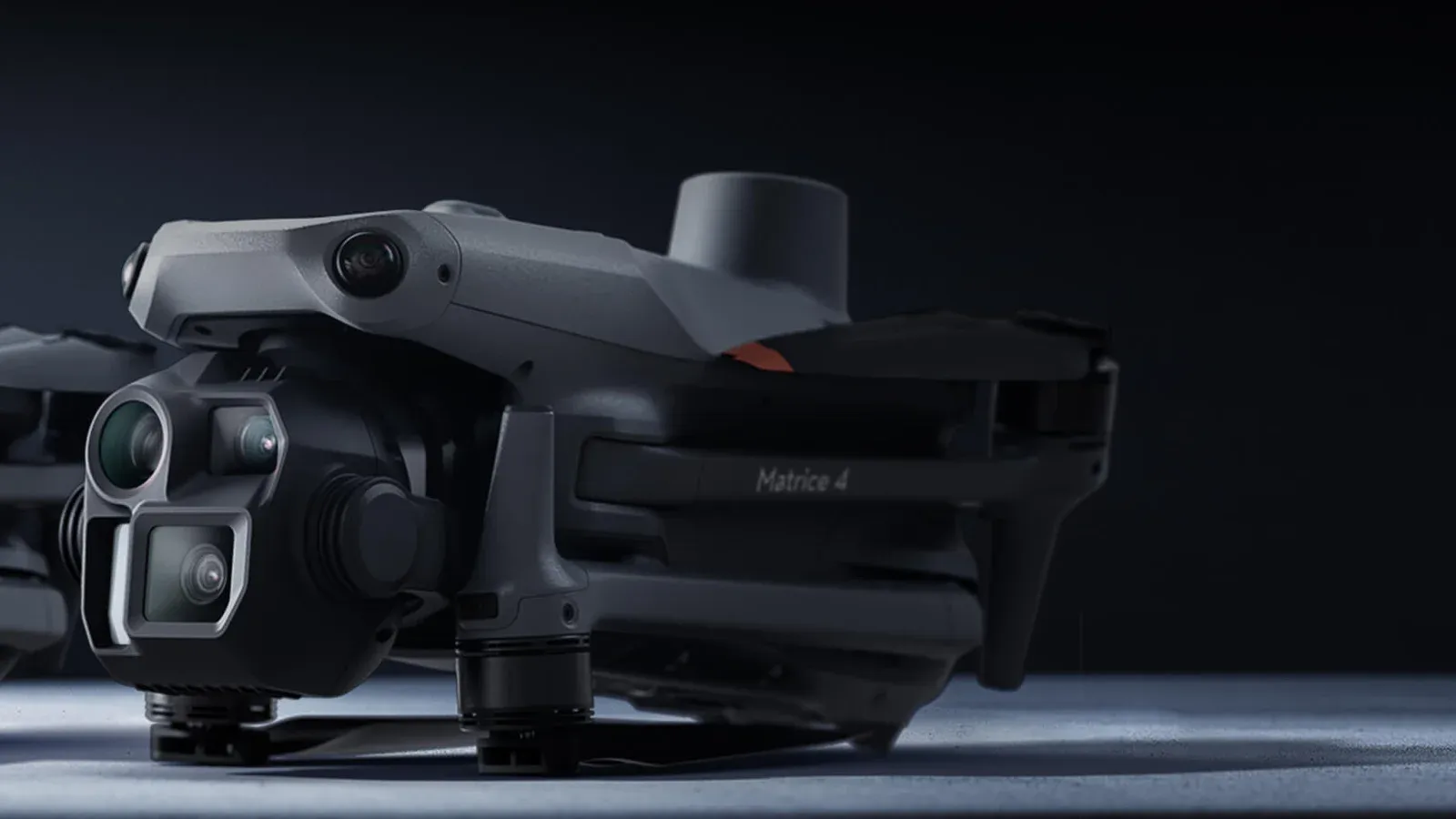 DJI Matrice 4E folded enterprise drone on a dark studio floor, its three-camera gimbal with the large square mapping lens at the front