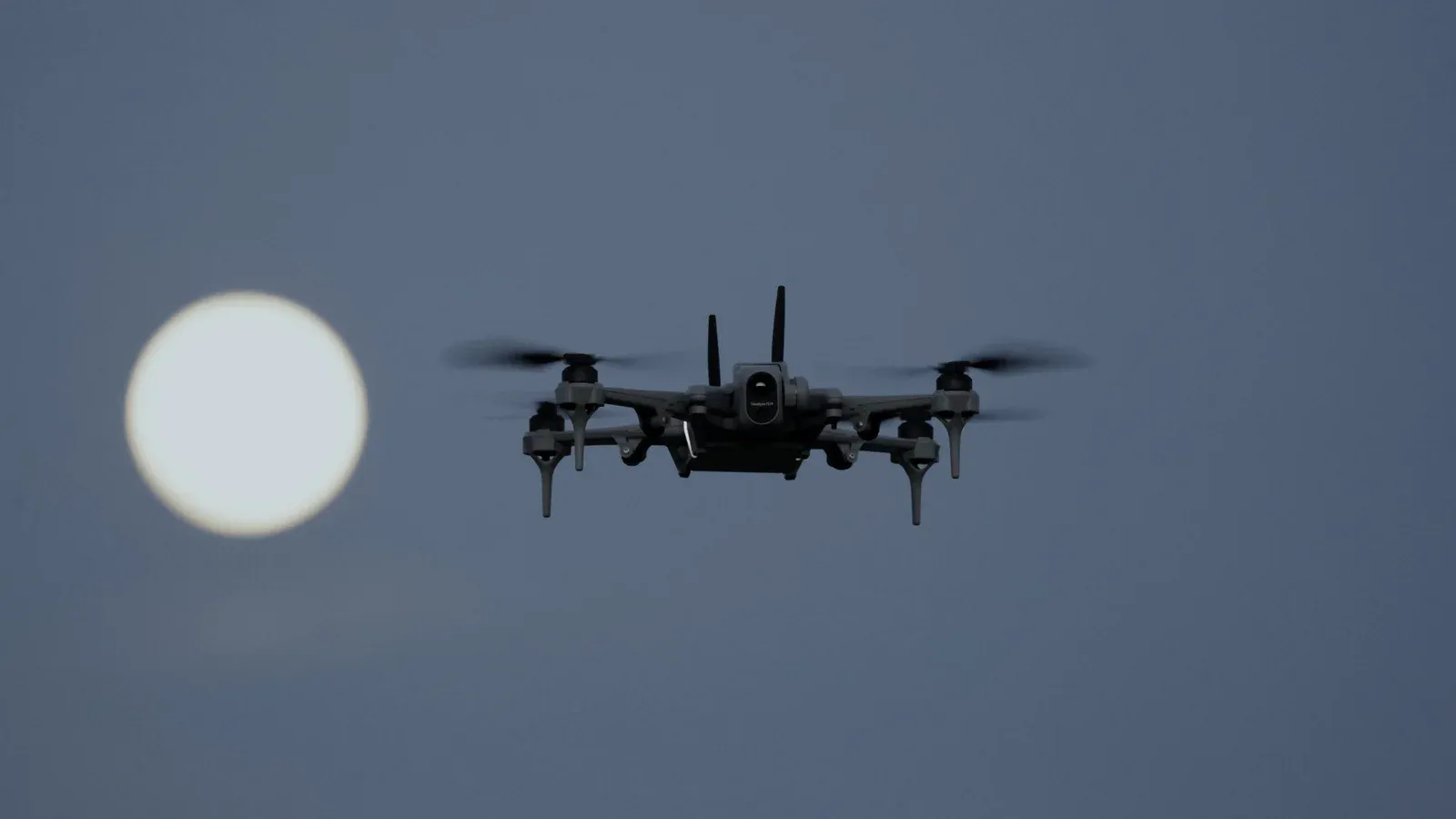 Teal 2 dark grey quadcopter with two antennas and a gimbal camera, hovering against a dusk sky with the full moon to its left