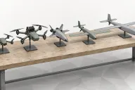 Illustration of five different generic drones lined up on a long workbench, from a small quadcopter to a fixed-wing aircraft, on a white background