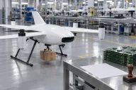 A fixed-wing delivery drone with a tethered package grounded on a factory floor beside a stack of printed circuit boards, a paper form and a rubber stamp on a steel table under bright overhead lights
