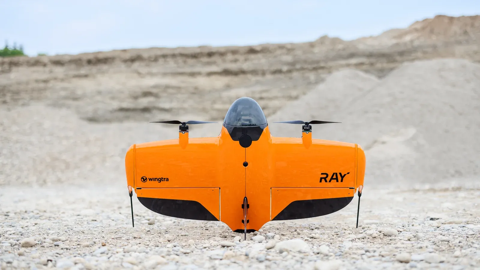 Orange WingtraRAY tailsitter drone standing upright on its tail on quarry gravel, wings spread, with two propellers along the top edge and spoil heaps behind
