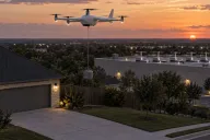 A white fixed-wing delivery drone with five rotors hovering above a suburban driveway at dusk, lowering a small pod on a tether, with a row of charging docks on a flat commercial rooftop in the background